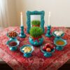 Turquoise Hamedan enamel Haft-sin set arranged on red Persian tapestry cloth with candles lit.