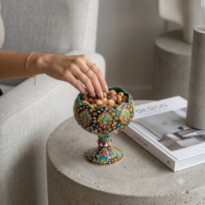 Hand-painted Hamedan enamel pottery nut bowl on concrete table with hand reaching for pistachios.