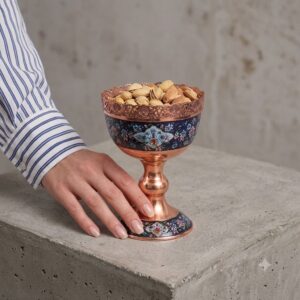 Hand holding a Persian handcrafted Copper and Pardaz nut bowl filled with pistachios on a concrete surface.