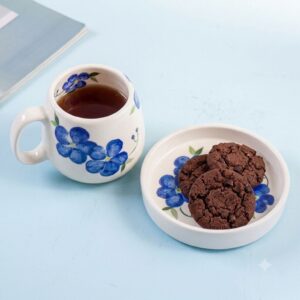 Handmade white ceramic mug with blue painted flowers alongside chocolate chip cookies on a matching saucer.