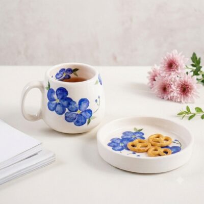 Sky Flower ceramic mug and pretzel-filled saucer displayed on a white tabletop next to pink flowers.
