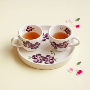 Top-down view of two violet floral ceramic tea cups on a matching round tray with loose flower petals.