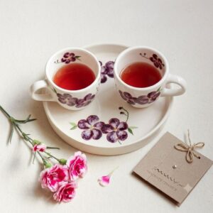 Violet ceramic tea set with artisan craft card and fresh pink flowers on a light linen surface.