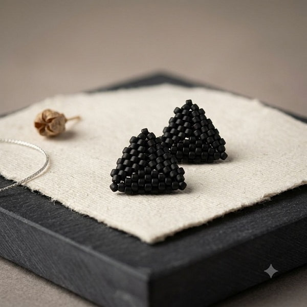 Pair of 3D woven matte black triangle beaded stud earrings displayed on textured artisan paper.
