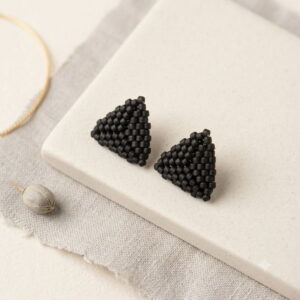 Flat lay of matte black 3D triangle beaded earrings on cream ceramic tile with textured linen fabric.