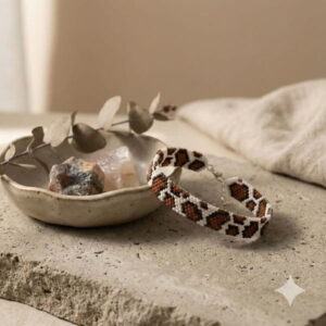 A handmade brown and white cow print beaded bracelet resting next to a decorative ceramic bowl with natural crystals.