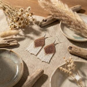 Elegant caramel-toned beaded fringe earrings laid out on a rustic fabric background surrounded by dried flowers and natural driftwood.