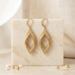 Hand-woven gold-toned seed bead diamond earrings on ivory marble pedestal.