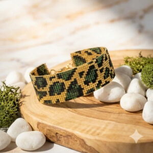 Handmade green leopard print beaded cuff bracelet displayed on a wooden platter surrounded by white pebbles.