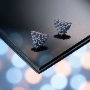 Periwinkle triangle stud earrings resting on reflective dark glass surface with bokeh lighting.