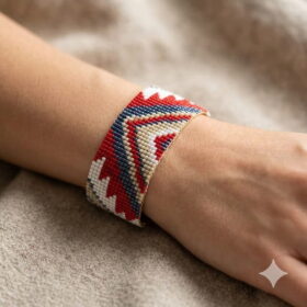 Detailed side-angle shot of a red and gold-toned geometric pattern beaded artisan cuff bracelet on a neutral texture.