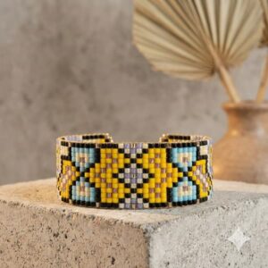 Close-up of a handmade yellow and blue mosaic pattern seed bead bracelet with gold-toned accents on a concrete block.
